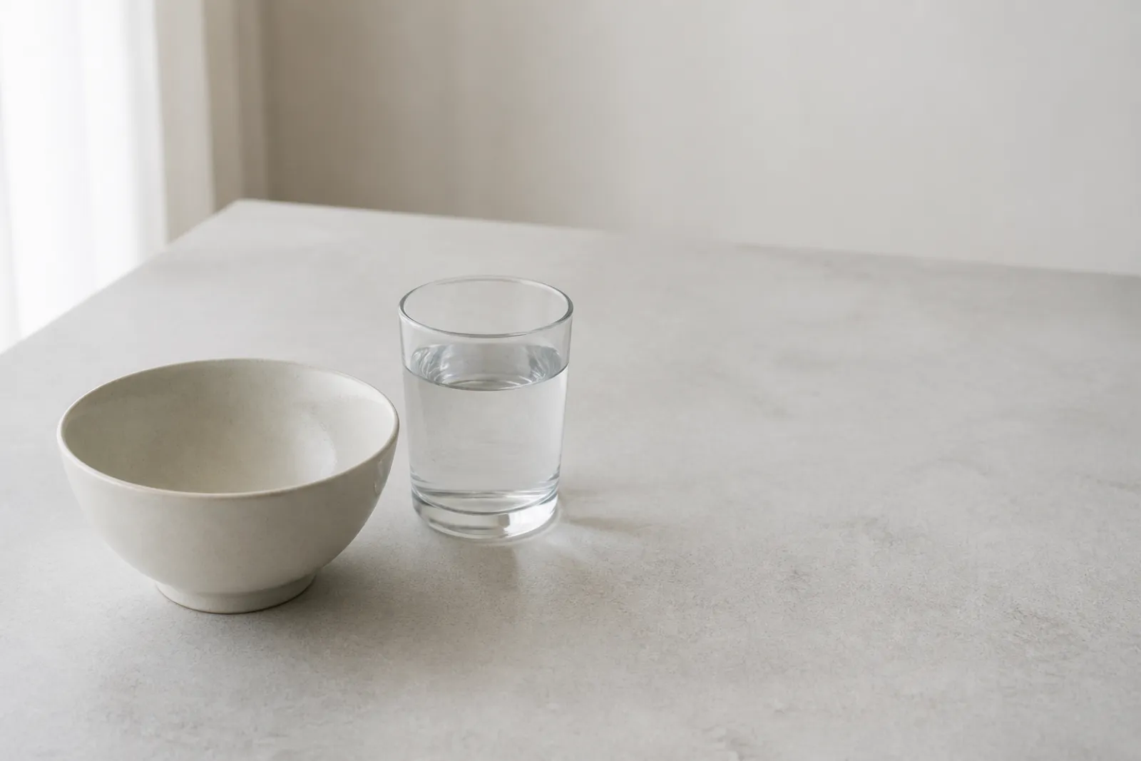 An empty ceramic bowl and a glass of water on a plain table in morning light.