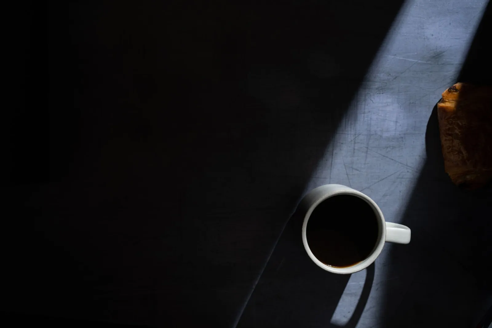 A coffee cup and a pastry on a flat surface in dramatic low-angle light.