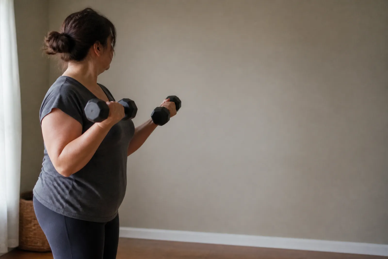A middle-aged woman lifting a pair of dumbbells in a plain room in flat daylight.