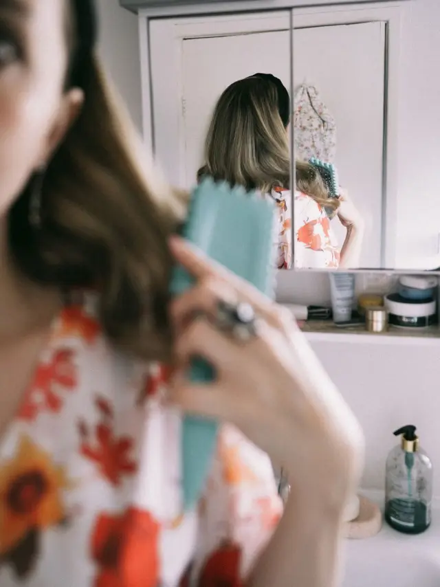 A woman in a floral dress brushing her hair in front of a mirror.