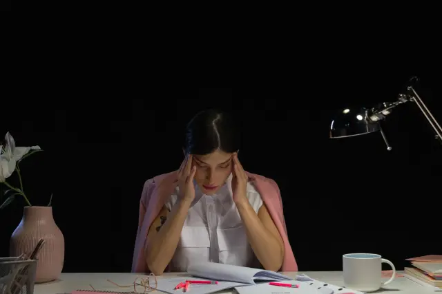 A woman at a desk with her hands pressed to her head, surrounded by papers.