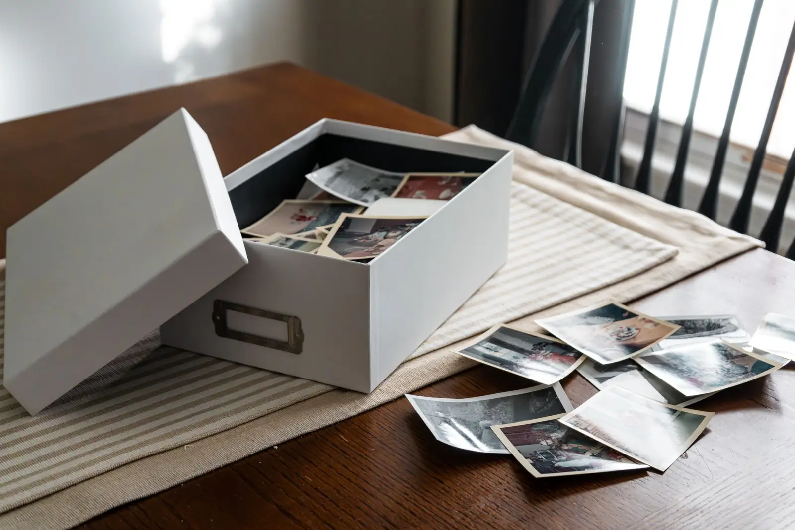 An open box of vintage family photographs on a wooden table beside a window.