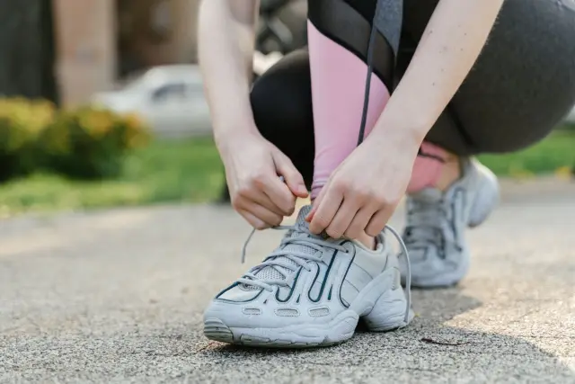 A woman crouched down tying the laces of her trainers on a park path in morning light.