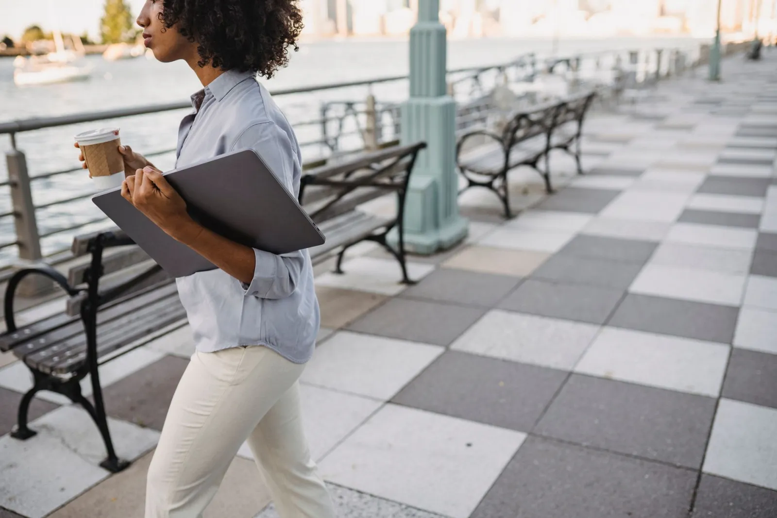 A woman walking along a riverside promenade in the morning, holding a coffee cup.