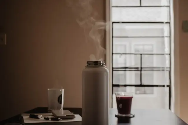 A steaming flask and warm drink on a table by a bright window.