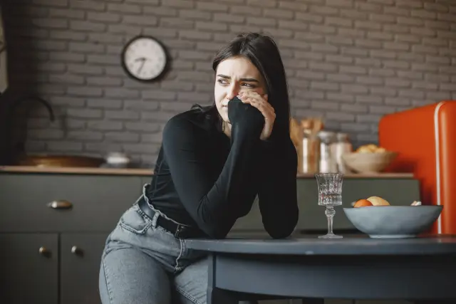 A woman sitting indoors in a pensive mood, hands resting in her lap.