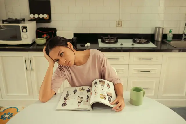 A woman sitting alone at a kitchen table with a cup of coffee, lost in thought.