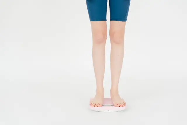 Crop anonymous female in sportswear measuring weight on scales for body control against white background.