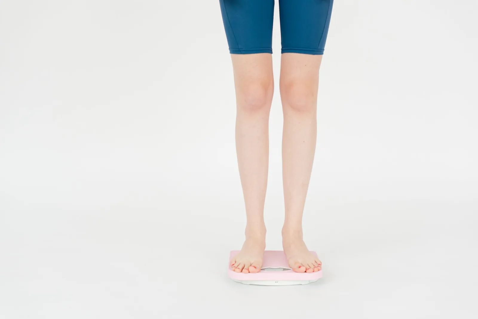 Crop anonymous female in sportswear measuring weight on scales for body control against white background.