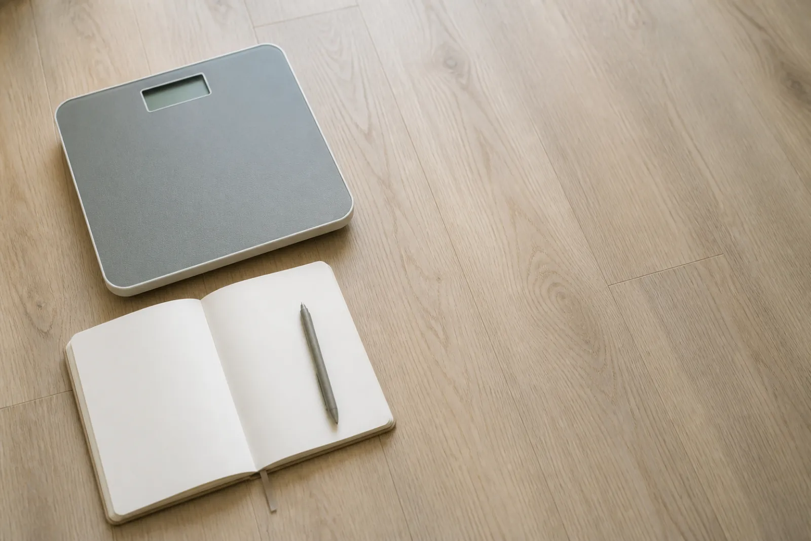 A bathroom scale beside an open notebook on a plain floor in flat daylight.
