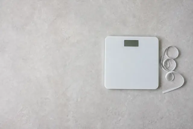 A floor scale and a coiled tape measure resting side by side on a plain bathroom floor.