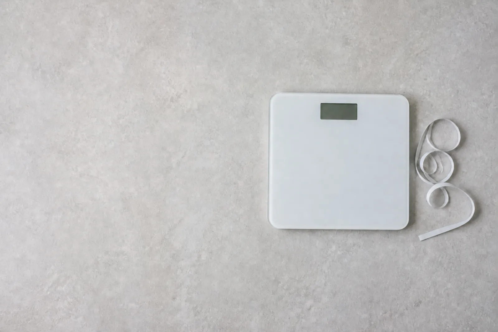 A floor scale and a coiled tape measure resting side by side on a plain bathroom floor.