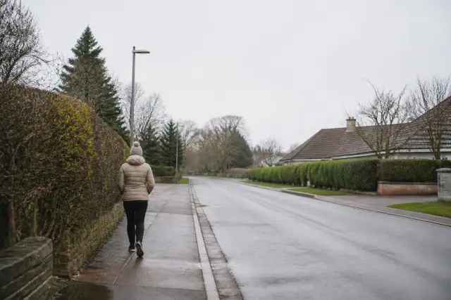 A person walking along a suburban pavement in flat overcast daylight.
