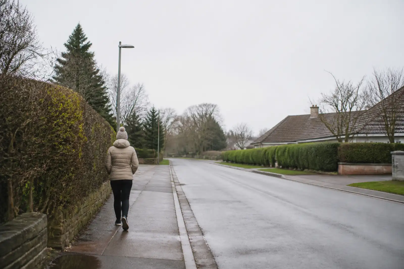 A person walking along a suburban pavement in flat overcast daylight.