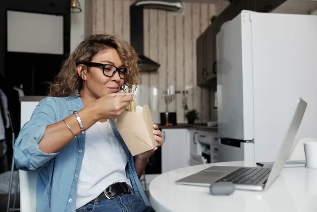 A woman eating a takeaway meal quickly while working at a laptop.