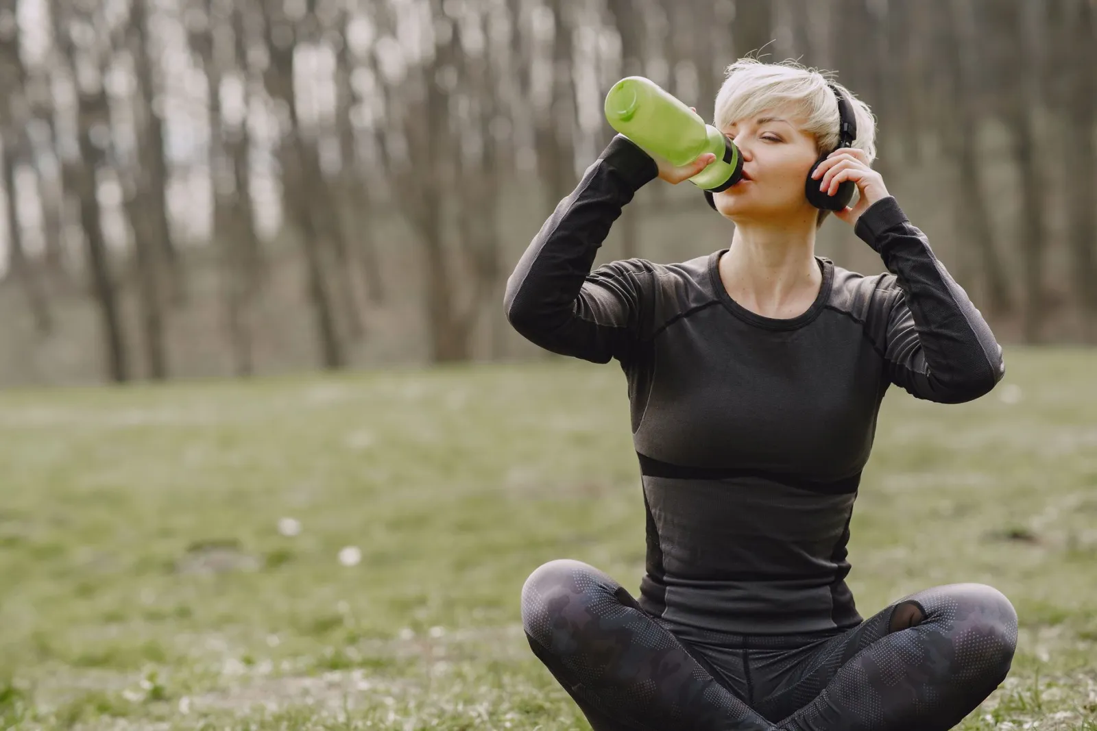 A woman in activewear drinking water while sitting outdoors.