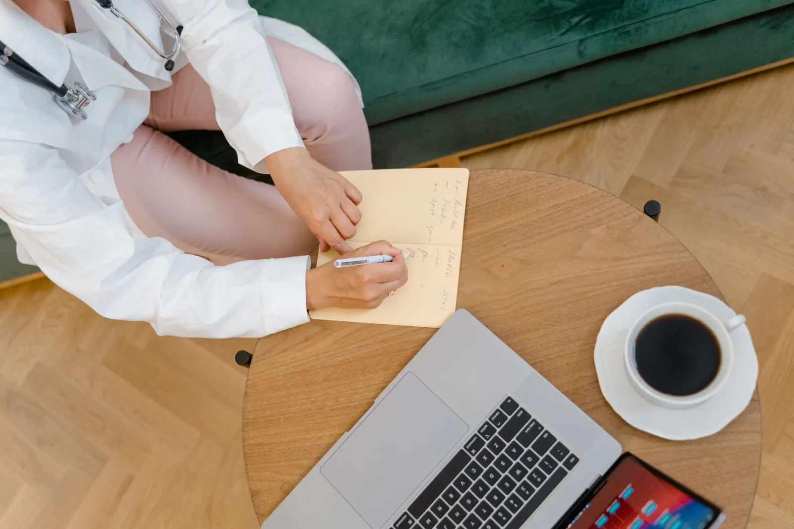 Overhead view of a doctor writing notes at a desk with a laptop and a cup of coffee.