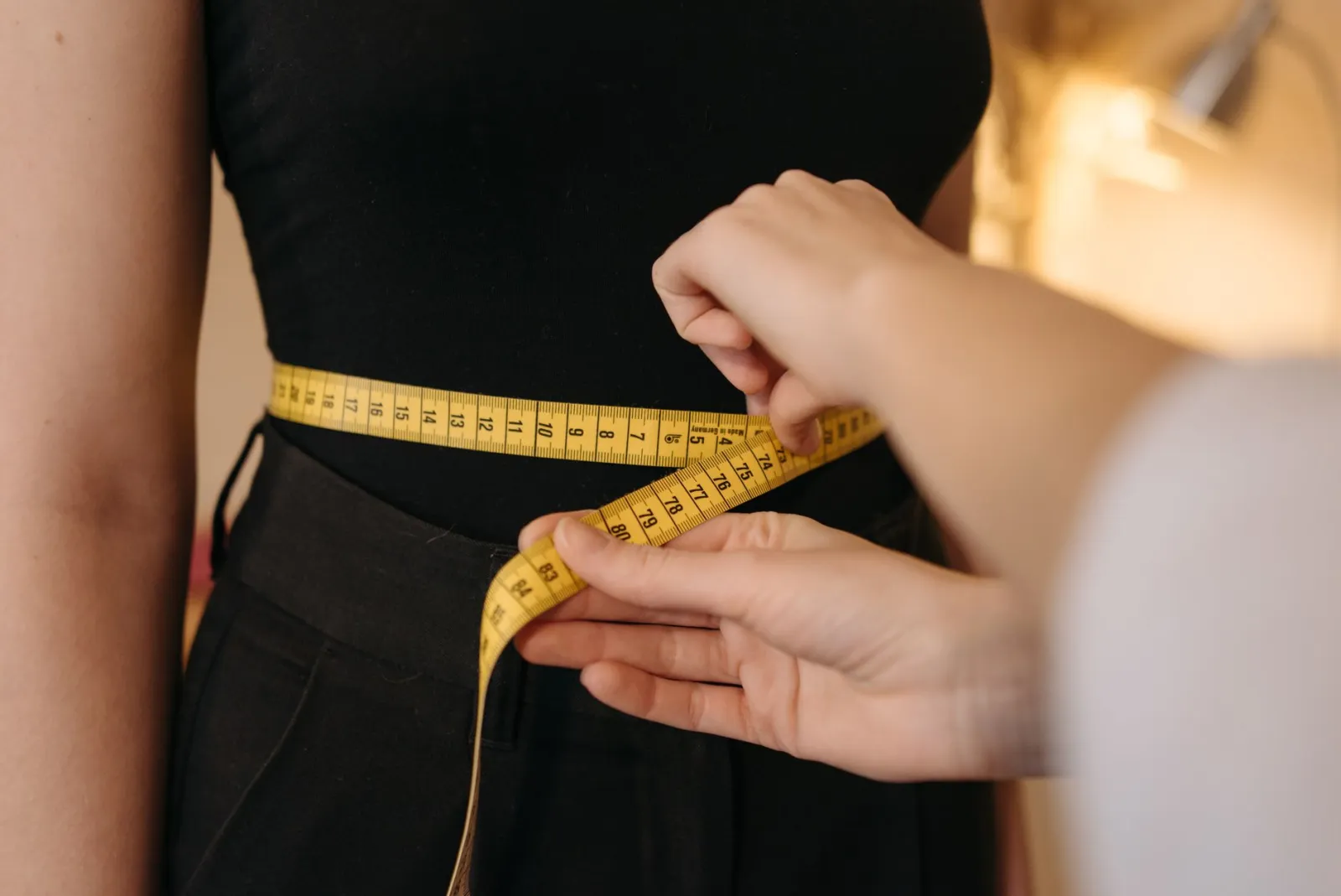 A tailor's hands measuring a person's waist with a yellow tape measure, cropped close with no face visible.