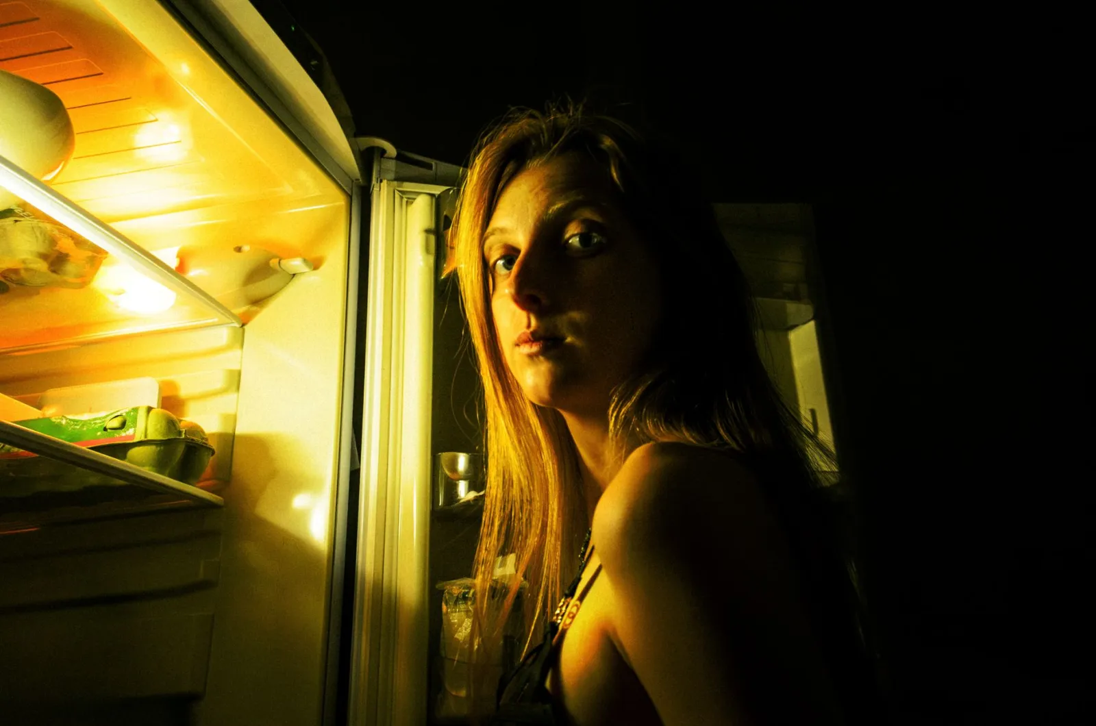 A person standing in a dim kitchen at night, looking thoughtfully into an open refrigerator.