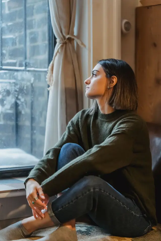 A woman sitting cross-legged by a window, looking out thoughtfully.