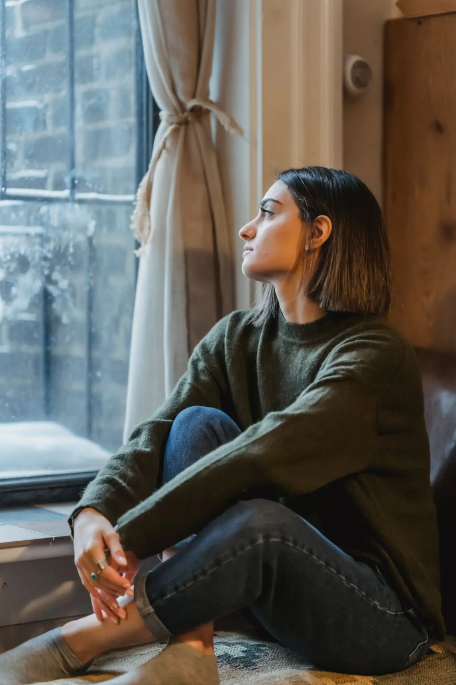 A woman sitting cross-legged by a window, looking out thoughtfully.