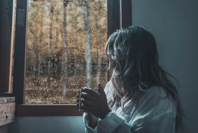 A woman holding a warm mug in both hands, gazing out through a rain-speckled window.