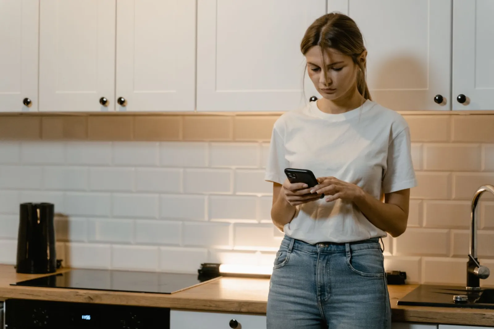 A young woman peacefully holding her smartphone in a modern kitchen, looking thoughtful.