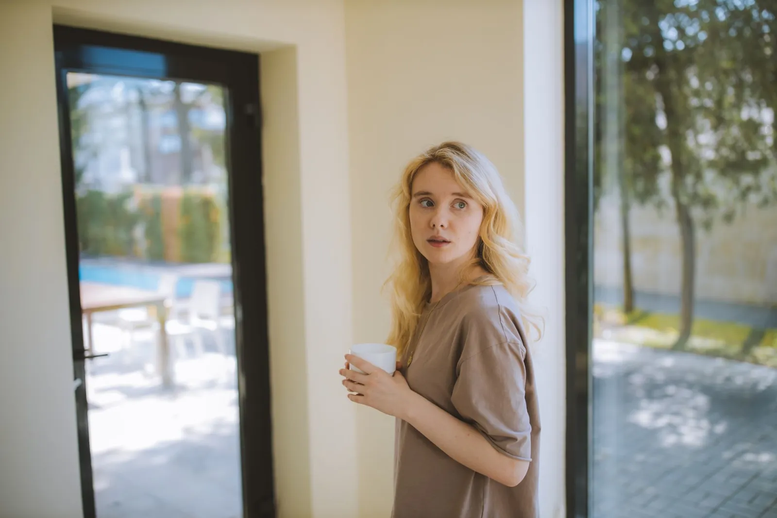 A woman holding a warm cup by tall glass doors, looking back into the room.